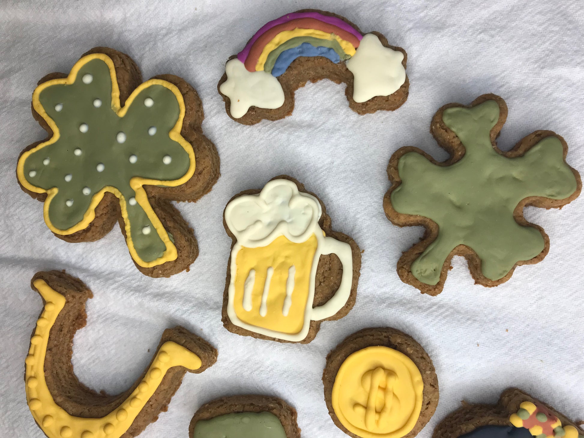 Decorative cookies shaped like a rainbow, clover, beer mug, and dollar sign on a white surface.