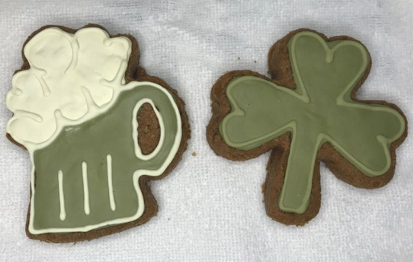 Two cookies shaped like a mug and a clover with green icing on a white surface.
