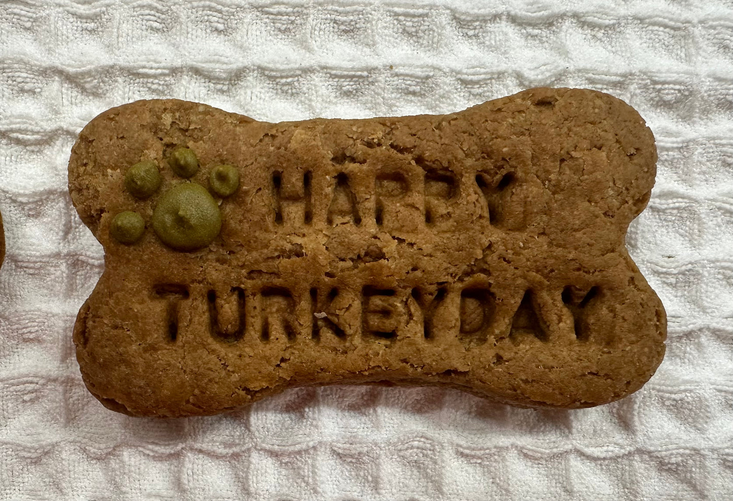 Bone-shaped dog treat with the words ‘Happy Turkey Day’ stamped on it and a small green paw print design, placed on a textured white cloth background.