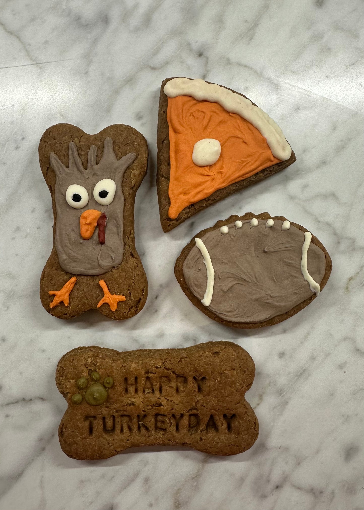 Assortment of Thanksgiving-themed dog treats on a marble surface, including a bone-shaped treat with a turkey face, a slice of pumpkin pie, a football, and a bone with ‘Happy Turkey Day’ stamped on it.