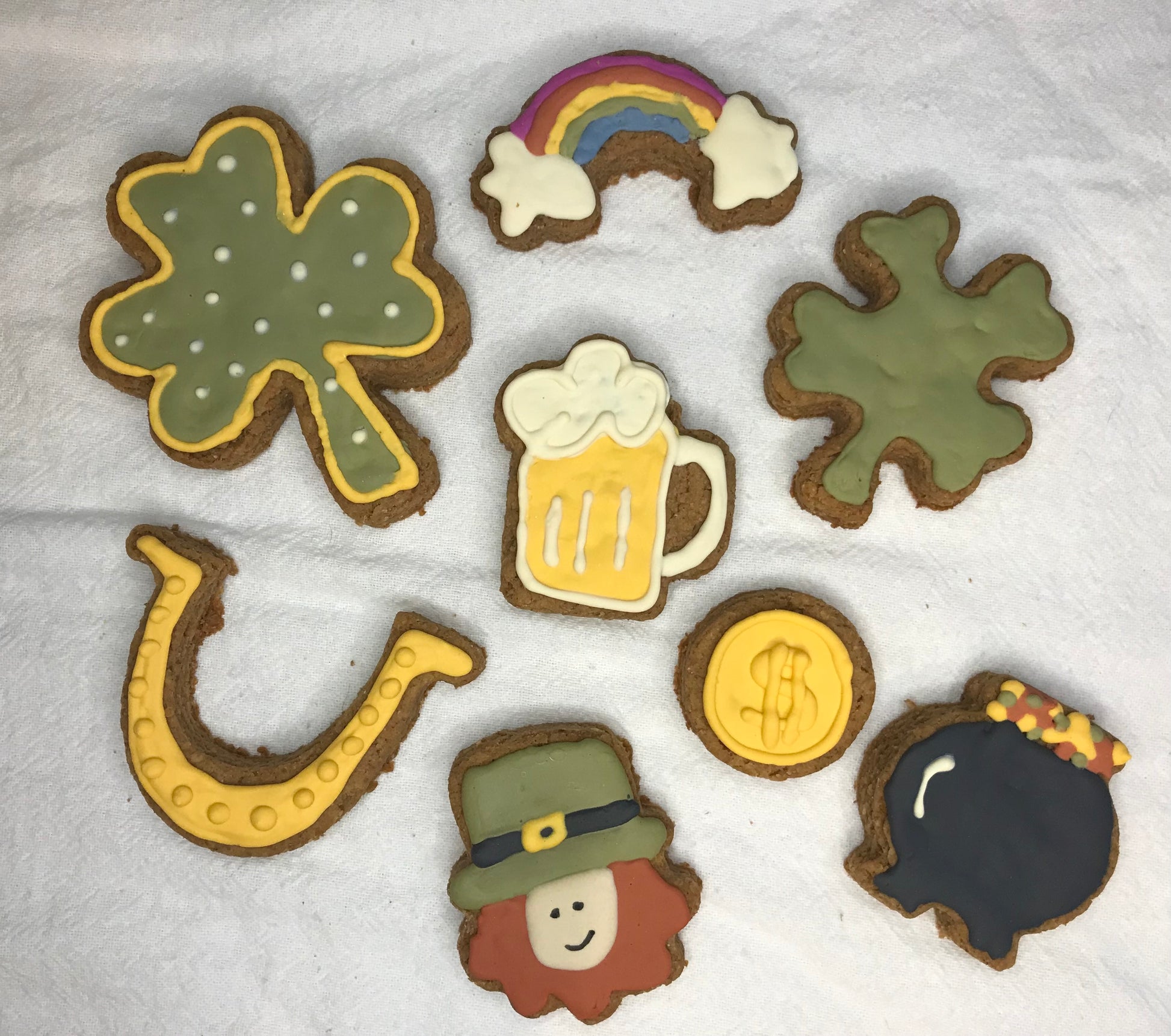 Decorative cookies shaped like a four-leaf clover, rainbow, beer mug, gold coin, leprechaun, and pot of gold on a white surface.