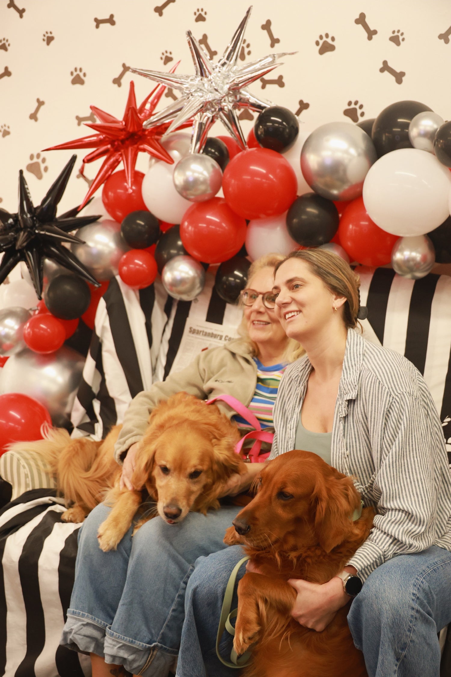 Two women with dogs in front of a festive backdrop with balloons and decorations.